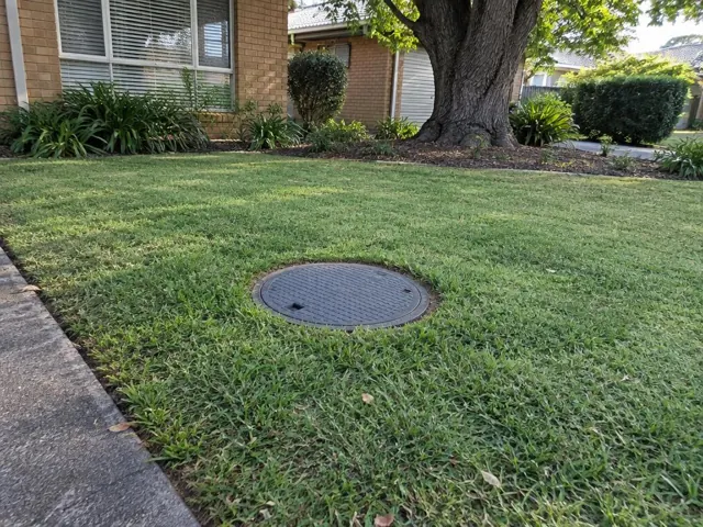 Round capped sewer access cover set in a neat lawn beside a brick house in Caringbah